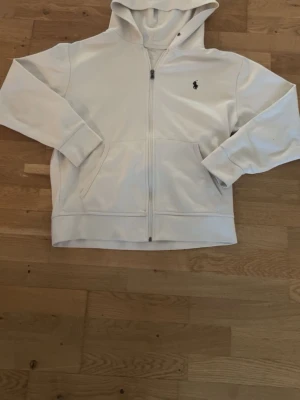 Vit zip hoodie från Polo Ralph Lauren - Snygg vit hoodie med dragkedja från Polo Ralph Lauren. Klassisk design med huva, två fickor framtill och den ikoniska lilla loggan broderad på bröstet. Perfekt för en clean och stilren look. Tillverkad i mjukt material för extra komfort.