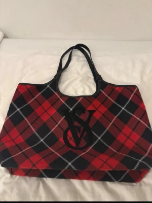 Viktoria s Sekrets Rutig shoppingväska i rött och svart - Stor shoppingväska med rutigt mönster i rött, svart och vitt. Väskan har svarta handtag och ett stort broderat monogram på framsidan. Materialet ser ut att vara tyg med en mjuk känsla och väskan har en rymlig form som passar till mycket.