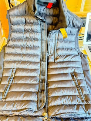 Svart dunväst med Parajumpers  - Snygg svart dunväst med horisontella sömmar och två dragkedjeförsedda fickor framtill. Västen har hög krage och en gul detalj vid bröstet. Perfekt för lager-på-lager och håller dig varm under kyliga dagar.