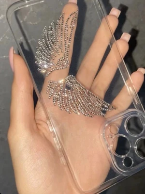 Genomskinligt mobilskal med strassvingar - Snyggt genomskinligt mobilskal dekorerat med glittriga strass i form av änglavingar. Skalets material är plast och det har öppningar för kamera och knappar. Perfekt för dig som vill ha ett unikt och trendigt skydd till mobilen.