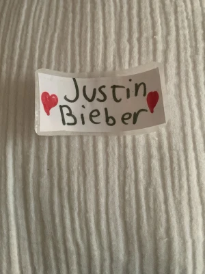 Handgjord Justin Bieber skylt - En liten handgjord klistermärke med texten 'Justin Bieber' skrivet i svart och dekorerad med två röda hjärtan. Klistermärken är gjord av papper och tejp. Perfekt för fans eller dekoration.
