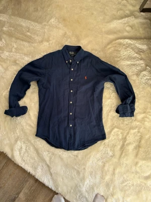 Marinblå skjorta från Polo Ralph Lauren - Snygg marinblå skjorta från Polo Ralph Lauren i slim fit-modell. Skjortan har klassisk krage, knappar framtill och den ikoniska broderade loggan på bröstet. Perfekt för en stilren look.
