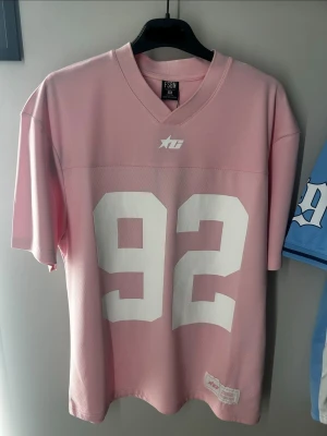 Rosa mesh jersey med nummer 92 - Säljer en rosa sportig mesh jersey med stort vitt nummer 92 på framsidan och v-ringad hals. Tröjan har korta ärmar och är tillverkad i ett lätt och ventilerande material, perfekt för träning eller streetwear.många storlekar