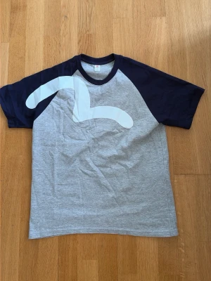 Grå och marinblå t-shirt med tryck - Snygg t-shirt i grått med marinblå ärmar och krage. Framsidan har ett stort vitt grafiskt tryck. Klassisk raglanmodell med korta ärmar och rund hals. Perfekt för en avslappnad stil.