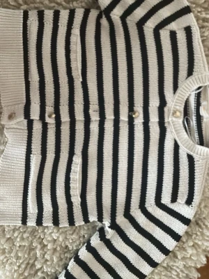 Randig stickad kofta med knappar - Snygg stickad kofta med svarta och vita horisontella ränder. Koftan har rund hals, dekorativa guldknappar framtill och två små fickor. Perfekt att slänga över en t-shirt för en chill look.