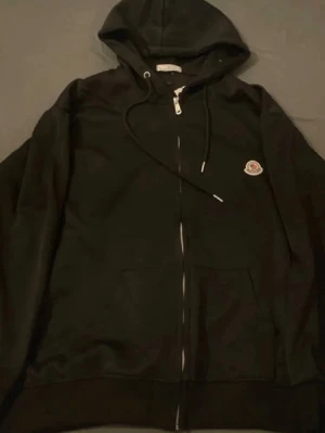 Svart hoodie med dragkedja och logga - Säljer en svart hoodie med dragkedja och huva. Den har en liten broderad logga på bröstet och praktiska fickor framtill. Klassisk modell med snörning i huvan och ribbade muddar. Perfekt för en chill och stilren look.