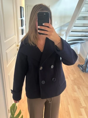 Mörkblå kappa - Stilren mörkblå skepparkavaj med dubbelknäppning och bred krage. Jackan har två framfickor och uppvikta ärmslut för en klassisk look. Perfekt för dig som vill ha en tidlös och snygg jacka till höst och vinter.