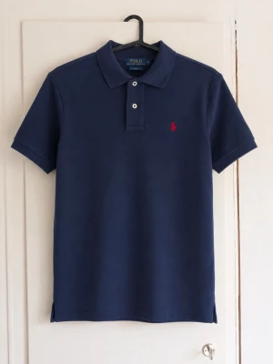 Ralph Lauren piké - Säljer en klassisk marinblå pikétröja från Polo Ralph Lauren med den ikoniska röda loggan på bröstet. En tidlös modell som passar till både vardag och finare tillfällen. ✔️ Broderad Polo-logga ✔️ Stilren marinblå färg ✔️ Bekväm passform ✔️ Enkel att matcha med jeans eller chinos Storlek: L (14-16 år) Skick: 8/10