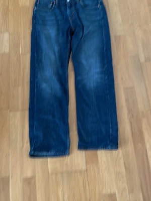 Mörkblå raka Levis jeans - Storlek 31/32 Säljer ett par mörkblå jeans med rak passform och klassisk femficksdesign. Jeansen har en lätt tvättad look och är tillverkade i denim. Perfekta för dig som gillar en avslappnad stil.
