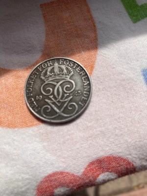 Svensk mynt 1943 med krona - Säljer ett svenskt mynt från 1943 med texten 'Med folket för fosterlandet' och en kunglig krona ovanpå ett monogram. Myntet är runt och i silverfärgat metallmaterial. Perfekt för samlare av äldre svenska mynt.