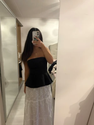 Svart axelbandslös peplum topp - Snygg svart axelbandslös topp med peplumskärning som ger en markerad midja och volang nertill. Toppen är tight upptill och har en stilren, elegant look. Perfekt att styla med kjol eller jeans för en trendig outfit.