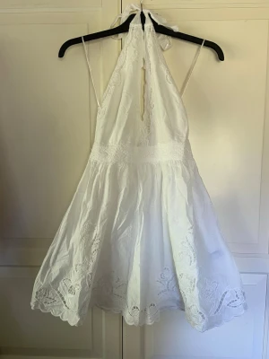 Vit kort klänning med spetsdetaljer - Söt vit kortklänning med halterneck och knyt i nacken. Klänningen har fina spetsdetaljer och broderade mönster längs fållen. Perfekt för varma dagar och somriga tillfällen. Lätt och luftig modell med omlottkänsla och markerad midja.