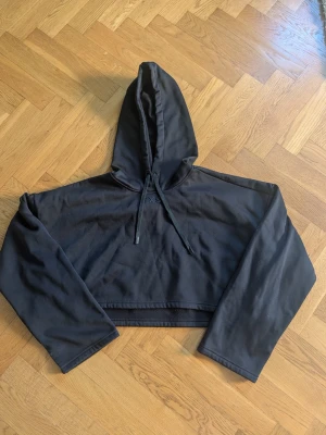 Svart croppad hoodie med huva - En svart croppad hoodie med huva och dragsko. Hoodien har långa ärmar och en enkel, stilren design utan synliga loggor eller tryck. Perfekt för en avslappnad och trendig look. Materialet ser ut att vara mjukt och bekvämt, troligen bomullsblandning.