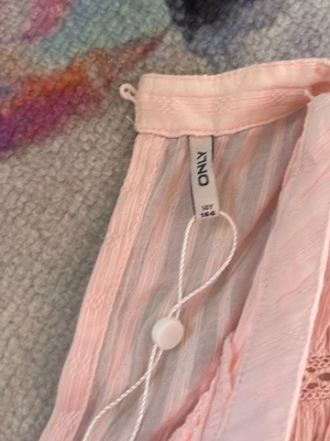Ljusrosa topp från ONLY - Söt ljusrosa topp från ONLY i ett tunt, lätt material med diskret struktur. Toppen har fina detaljer som smock i midjan och spets längs kanterna. Perfekt för sommarens alla tillfällen!