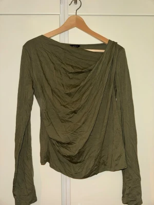 Olivgrön draperad topp från Lindex - Snygg olivgrön topp från Lindex med draperad front och lång ärm. Toppen har en asymmetrisk halsringning och är tillverkad i ett mjukt, tunt material som ger ett avslappnat intryck. Perfekt för dig som gillar unika detaljer och stilren design.