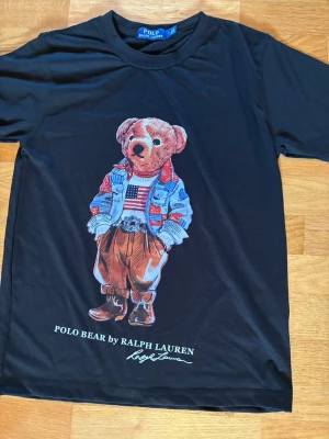 Svart Polo Bear t-shirt Ralph Lauren - Snygg svart t-shirt från Ralph Lauren med ikoniskt Polo Bear-tryck framtill. Björnen har på sig jeansjacka, randig tröja och bruna byxor. T-shirten har rund hals och korta ärmar. Perfekt för dig som gillar streetwear och klassiska prints.     Kontakta gärna vid frågor och prisförslag. Skickar inom 24h.