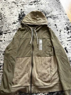 Olivgrön hoodie från Hugo Boss - Säljer en olivgrön hoodie från Hugo Boss med dragkedja och vita snören i huvan. Hoodien har en broderad logga på bröstet och är tillverkad i mjukt bomullsmaterial. Tvådelad färg med mörkare och ljusare olivgrönt. Perfekt för en avslappnad stil.