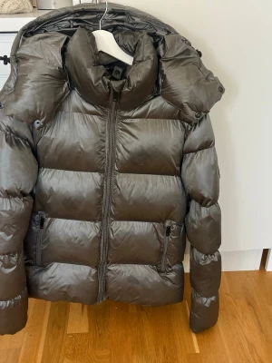 Mörkgrå/olivgrön dunjacka med huva - Snygg mörkgrå/olivgrön dunjacka med glansig finish och stor, vadderad huva. Jackan har två dragkedjeförsedda fickor framtill och breda, quiltade paneler som ger en puffig look. Perfekt för kalla dagar och riktigt varm. Passar både dam o herr. 