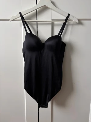 Svart body med vadderade kupor - Snygg svart body med vadderade kupor och justerbara axelband. Tillverkad i ett stretchigt, slätt material som ger en smickrande passform. Bodyn har en klassisk skärning och knäppning i grenen. Perfekt för att skapa en stilren look. Från märket Dorina/ Zalando. Använd en gång, nypris 329kr.