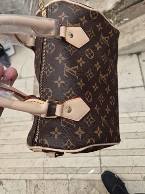 Brun  handväska - Snygg brun handväska från  med klassiskt monogrammönster i beige och guldiga detaljer. Väskan har två korta handtag i ljust läder och dragkedja upptill. Perfekt för dig som gillar lyxiga accessoarer och tidlös design. Tar ej emot bud 