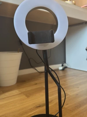 Ringlampa med mobilhållare - Säljer en ringlampa med stativ och mobilhållare. Lampan är vit och svart, har rund form och står på en stabil rund fot. Perfekt för streaming, videosamtal eller sminkning. USB-anslutning för enkel användning.
