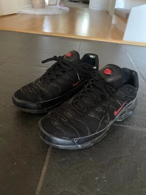 Svarta Nike TN sneakers - Säljer ett par svarta Nike TN sneakers med mesh- och syntetmaterial, röda detaljer och klassisk swoosh på sidan. Skorna har en chunky sula och lågt skaft för en sportig vibe. Perfekta för dig som gillar streetwear och vill sticka ut.