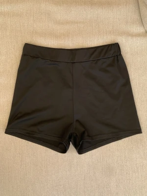 Svarta tighta shorts i syntet - Säljer ett par svarta, tighta shorts med hög midja. De är gjorda i ett stretchigt syntetmaterial som passar perfekt till träning eller dans. Enkel och stilren design utan synliga detaljer eller mönster.