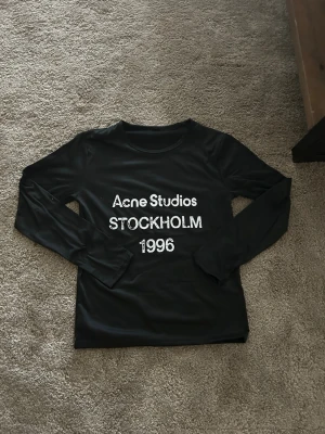 Svart långärmad topp Acne Studios - Svart långärmad topp från Acne Studios med tryck i vitt framtill där det står 'Acne Studios STOCKHOLM 1996'. Tunn och mjuk känsla, perfekt till jeans eller kjol för en clean look.