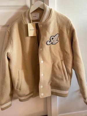 Axel Arigato Signature Bomber Beige College Jackets XS - Nypris 4000kr. Lappen finns kvar, aldrig använd. XS, känns som M. Snygg beige bomberjacka från Axel Arigato i modellen Signature Bomber. Jackan har ribbade muddar och krage med vita ränder, tryckknappar framtill och två fickor. Materialet är mjukt och teddy-liknande. Stor broderad logga på bröstet ger en cool streetwear-vibe. 