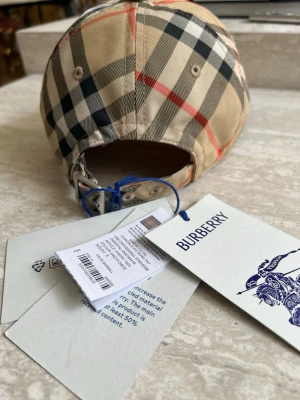 Beige keps med rutigt mönster Burberry - Snygg keps från Burberry i klassiskt beige rutigt mönster med svarta, vita och röda detaljer. Justerbart spänne bak i metall och bomullsmaterial. Perfekt accessoar för en trendig och stilren look.