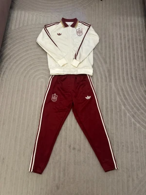 Vinrött och vitt träningsset Adidas - Snyggt träningsset från Adidas med vit zip-jacka och vinröda detaljer samt matchande vinröda byxor med vita ränder längs sidorna. Både jackan och byxorna har Adidas-logga och klubbmärke. Perfekt för sport eller chill. Materialet är troligtvis polyester.