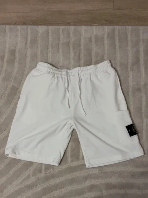 Vita stone island shorts  - Säljer ett par vita mjukisshorts med resår och dragsko i midjan. Shortsen har sidofickor och en ficka på benet med en svart patch med logga. Perfekta för en avslappnad och sportig stil.