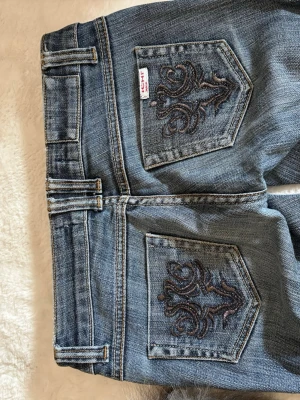 Blå jeans med broderade bakfickor - Säljer ett par blå jeans med snygga broderade detaljer på bakfickorna. Jeansen har låg midja och raka ben. Klassisk femficksmodell i slitstark denim, perfekta till vardagslooken. Passar dig som gillar en avslappnad men ändå trendig stil.