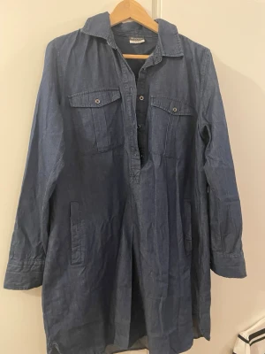 Blå jeansklänning med fickor - Säljer en blå jeansklänning med lång ärm, krage och knappar framtill. Klänningen har två bröstfickor med lock och knappar samt två sidofickor. Perfekt för en avslappnad och cool stil. (Är lite liten i stl så kanske som en 40.)
