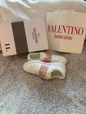 Valentino Garavani vita sneakers - Snygga vita sneakers från Valentino Garavani med bred rosa rem över vristen och nitar i silver. Skorna har en klassisk låg siluett och är tillverkade i slätt läder med vita snören och diskret logga på plösen och båda e vänster skor
