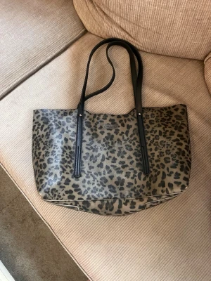 Leopardmönstrad handväska med svarta handtag - Cool handväska i leopardmönstrat konstläder med långa svarta handtag. Rymlig modell som rymmer allt du behöver. Perfekt för dig som vill sticka ut med ett djurmönster och gillar trendiga accessoarer.