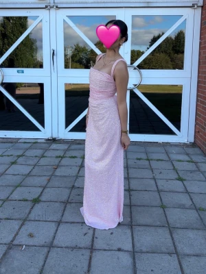 Ljusrosa glittrig långklänning - Elegant ljusrosa långklänning med smala axelband och glittrigt tyg. Klänningen har figurnära passform, snörning i ryggen och subtila volangdetaljer vid bysten. Perfekt för speciella tillfällen och student.