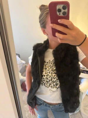 Svart fluffig pälsväst som skiftar i grönt - Säljer en svart, mjuk och fluffig pälsväst med öppen front.  Skiftar i grönt i ljus. Västen har en klassisk rak passform och är perfekt för att lyfta vilken outfit som helst. Materialet är fuskpäls som känns lyxigt och trendigt. Passar perfekt till jeans och t-shirt för en cool look. Det står ingen storlek men skulle säga att den passar som S eller M