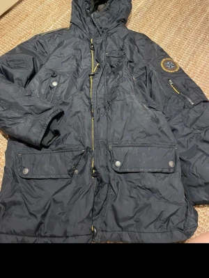 Mörkblå parkas med patch och huva - Snygg mörkblå parkas med huva och flera fickor, både med dragkedja och knappar. Jackan har en cool patch på ärmen och detaljer i gult vid dragkedjorna. Tillverkad i slitstarkt syntetmaterial, perfekt för kalla dagar.