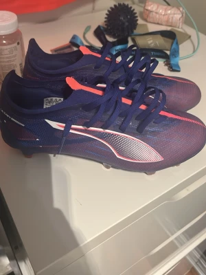 Blå och rosa fotbollsskor Puma - Säljer ett par snygga fotbollsskor från Puma i blått med rosa och vita detaljer. Skorna har snörning, låg profil och är tillverkade i ett lätt syntetmaterial för bra bollkontroll och grepp på planen. Perfekta för dig som vill sticka ut på matchen!