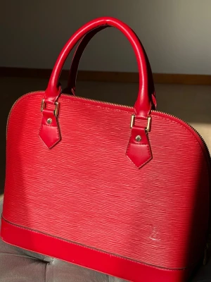 Röd strukturerad LV handväska i läder - Vintage Louis Vuitton Alma Epi i rött. Klassisk och tidlös Louis Vuitton Alma i Epi-läder med guldfärgade detaljer. Väskan har en strukturerad form, dubbla handtag och dragkedja.  Väskan är vintage och har tecken på användning som syns på bilderna. Insidan är i beige. Skriv gärna om du vill se fler bilder!