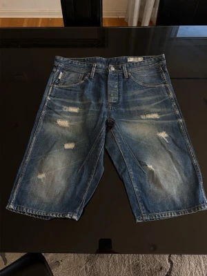 Jack & Jones denimshorts - Säljer ett par blå denimshorts med slitna detaljer och fem klassiska fickor. Shortsen har en loose passform och är tillverkade i bomull. Perfekta för en avslappnad stil under sommaren. 