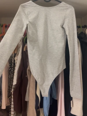 Grå långärmad body med djup rygg - Snygg grå body med långa ärmar och djup, rundad rygg. Tillverkad i mjukt stretchigt material som sitter tajt mot kroppen. Perfekt att styla med jeans eller kjol för en clean och enkel look.