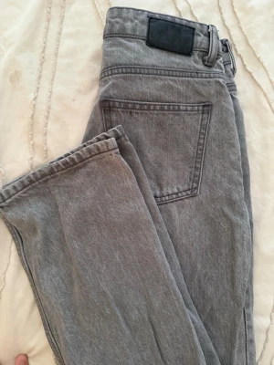 Grå raka jeans med fem fickor - Säljer ett par grå jeans med klassisk femficksmodell och raka ben. Jeansen har bälteshällor, knappgylf och en svart patch bak i midjan. Perfekta för en avslappnad och stilren look. Materialet är denim i bomull.