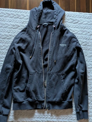 Balmain Paris Small Logo Zip-Up Hoodie - Svart huvtröja i premiumkvalitet med dragkedja och diskret Balmain-logotyp på bröstet.