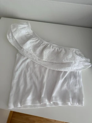 Vit oneshoulder topp med volang - Säljer en söt vit oneshoulder topp med bred volangkant och broderade detaljer. Toppen är ribbad och har en axel bar, vilket ger en trendig och somrig look. Perfekt till jeans eller kjol för en fräsch outfit.