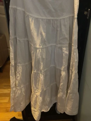 Vit långkjol i bomull med volanger - Säljer en vit långkjol i bomull med flera volanglager. Kjolen har en luftig och flowy känsla, perfekt för varmare dagar. Enkel och stilren design som är lätt att matcha med olika toppar. Knapp och dragkedja på sidan.