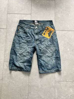 Blå jeansshorts från Karl Kani - Säljer ett par blå jeansshorts från Karl Kani med gul logga på framsidan och unika bruna detaljer på bakfickorna. Klassisk femficksmodell med knappgylf och normal passform. Perfekta för en avslappnad streetwear-look.