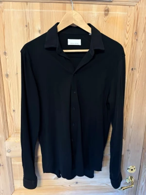 Svart skjorta från Casual Friday - Stilren svart skjorta från Casual Friday i klassisk modell med lång ärm och knappar framtill. Skjortan har en enkel krage och är tillverkad i ett mjukt material som känns skönt mot huden. Perfekt för en clean och modern look.