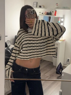 Randig croppad stickad tröja - Säljer en croppad stickad tröja i vitt med svarta ränder och diskret hålmönster. Tröjan har vida ärmar och avslappnad passform, perfekt för en trendig och chill look. Passar dig som gillar oversized och streetstyle.
